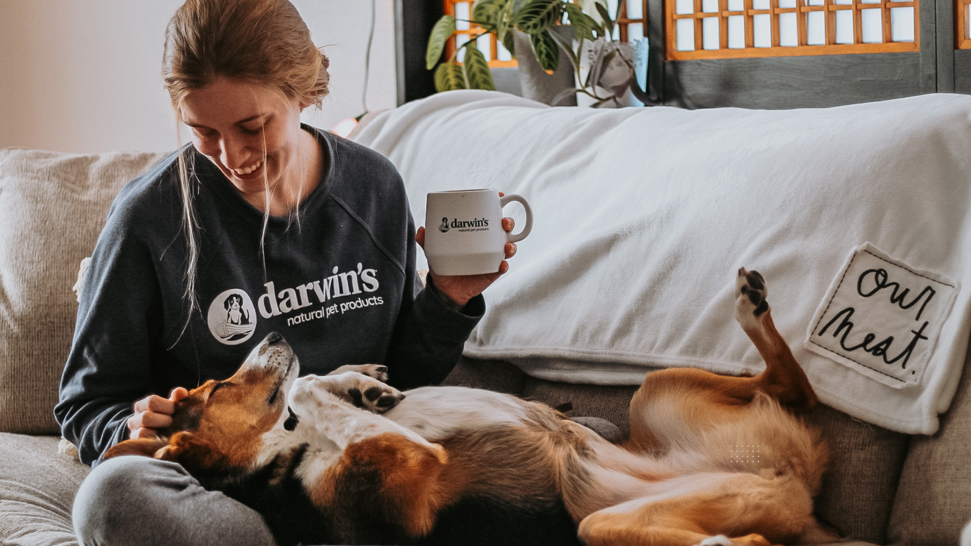 A dog sleeps while her owner looks down at the dog, happy. She is wearing a Darwin's raw dog food sweatshirt and drinking from a Darwin's mug.